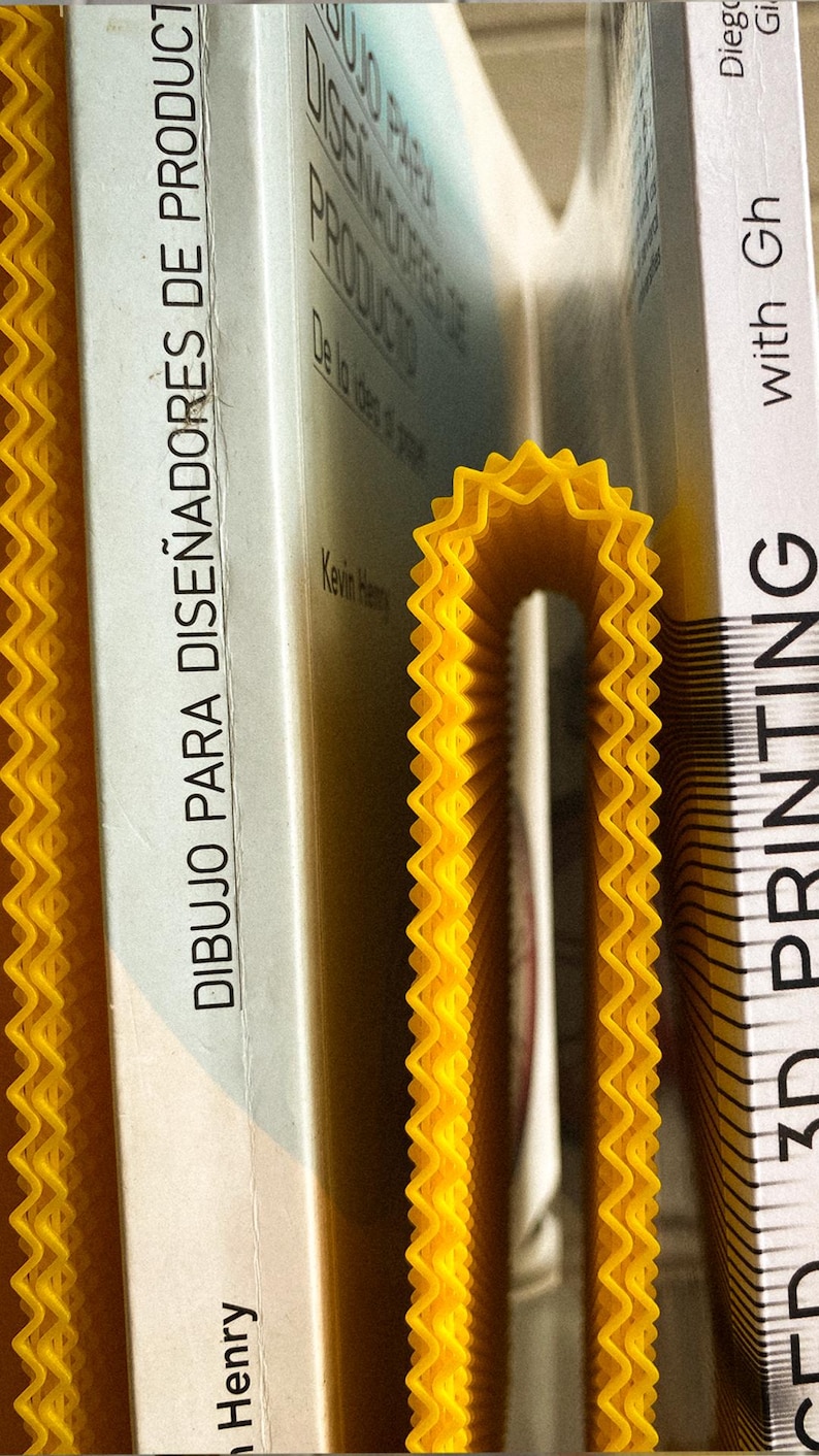 May include: A yellow, wavy-edged 3D-printed bookend supporting several books. The books have white covers with text in Spanish and English. The bookend's bright colour contrasts with the book covers, creating a visually appealing arrangement.