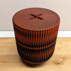 Puede incluir: Un contenedor cilíndrico impreso en 3D con una superficie texturizada. El contenedor es de color naranja oscuro con detalles negros. La parte superior del contenedor tiene una abertura en forma de cruz.