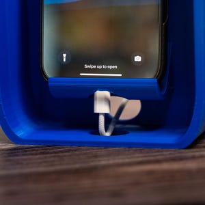 May include: A bright blue plastic smartphone charging dock with a rectangular frame. A smartphone is seated in the center, displaying the text Swipe up to open on the screen. A white charging cable is plugged into the bottom of the device.