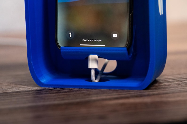 May include: A bright blue plastic smartphone charging dock with a rectangular frame. A smartphone is seated in the center, displaying the text Swipe up to open on the screen. A white charging cable is plugged into the bottom of the device.