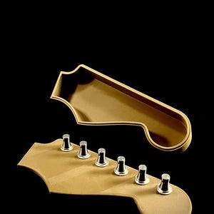 Puede incluir: Vista detallada de dos objetos con forma de clavijero de guitarra. El clavijero inferior tiene seis clavijas de afinaci&oacute;n. Ambos son de color marr&oacute;n claro con ribete dorado, sobre un fondo negro. El clavijero superior es una pieza separada.