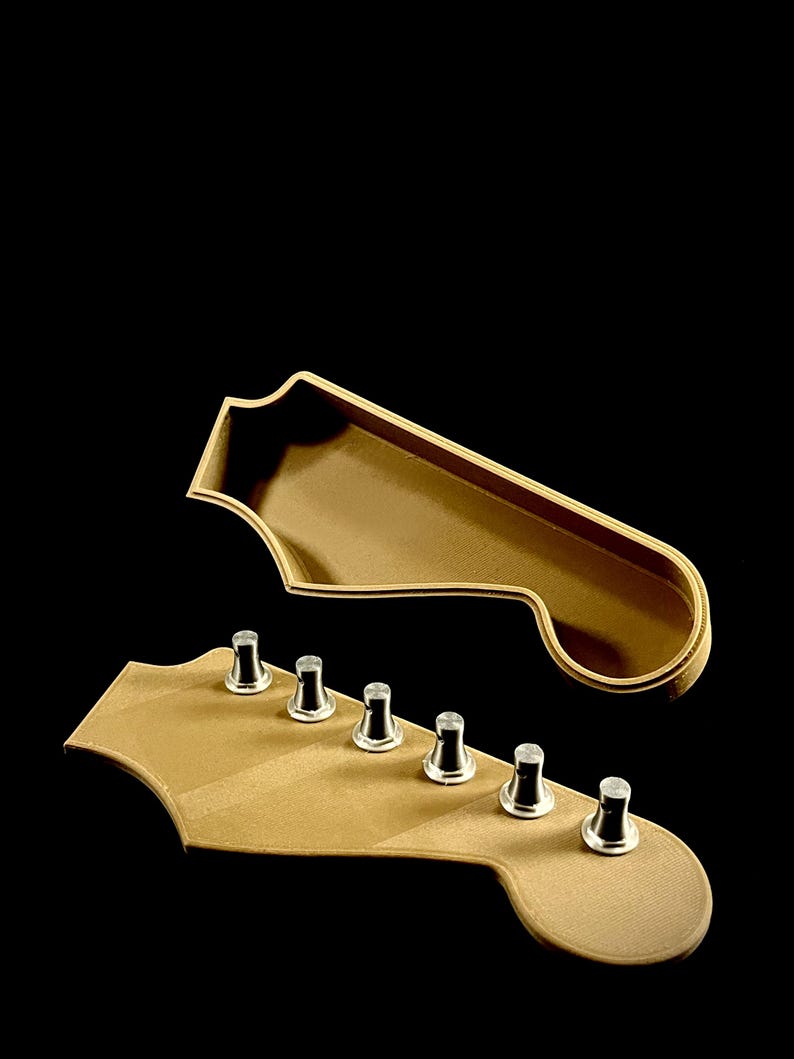 Puede incluir: Vista detallada de dos objetos con forma de clavijero de guitarra. El clavijero inferior tiene seis clavijas de afinaci&oacute;n. Ambos son de color marr&oacute;n claro con ribete dorado, sobre un fondo negro. El clavijero superior es una pieza separada.