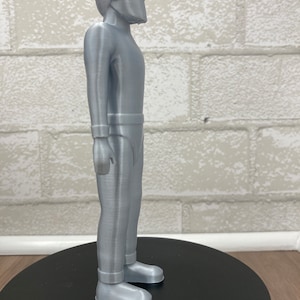 May include: A silver-colored 3D printed figurine of a person wearing a helmet and a jumpsuit. The figurine is standing on a black circular base.