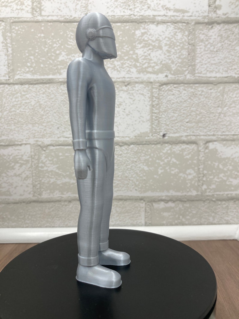May include: A silver-colored 3D printed figurine of a person wearing a helmet and a jumpsuit. The figurine is standing on a black circular base.