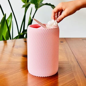 May include: A pink, textured wastebasket with a lid and a small wooden spoon inside. A hand is placing a crumpled tissue into the container. The wastebasket is cylindrical and sits on a wooden table.