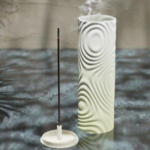 May include: White ceramic incense holder with a swirling pattern. The holder has a removable base and a single incense stick is burning inside.