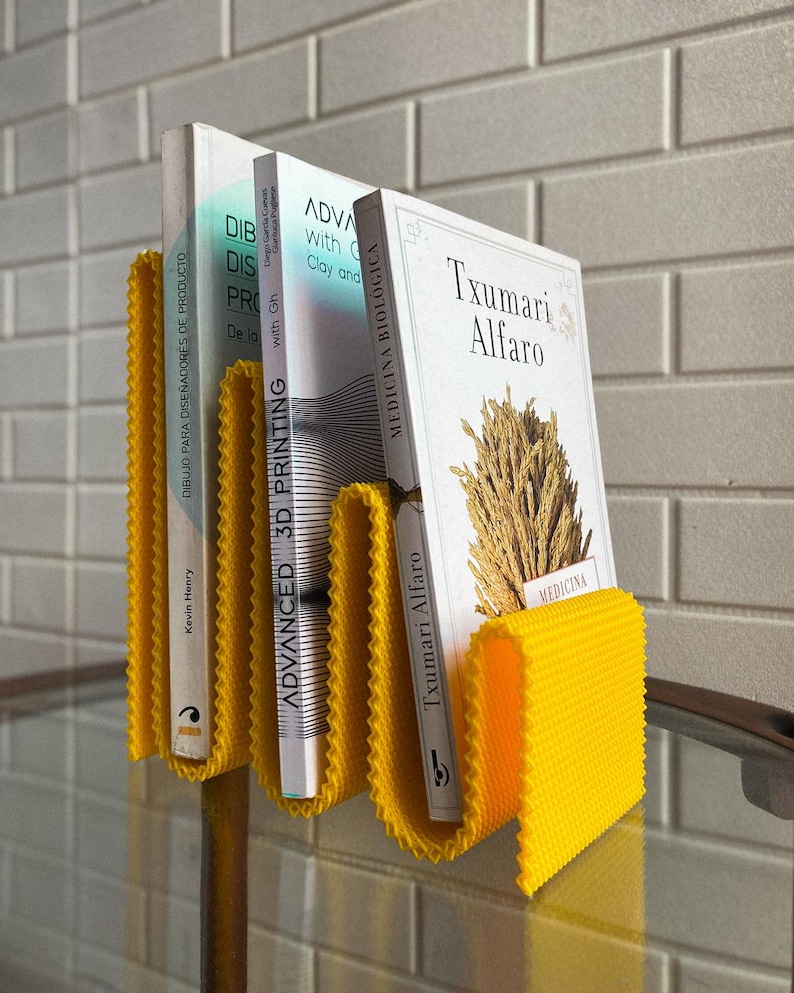 May include: A yellow, corrugated book stand holding three books. The books have white covers with text in English and Spanish. The stand is made of a textured material and is on a glass surface.