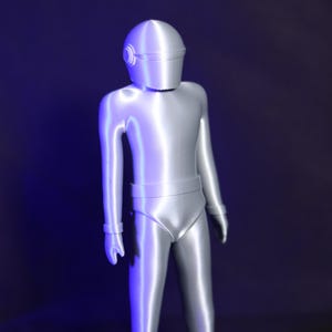 May include: A silver-colored robot figurine with a helmet and a smooth, shiny finish. The robot is standing upright with its arms at its sides.
