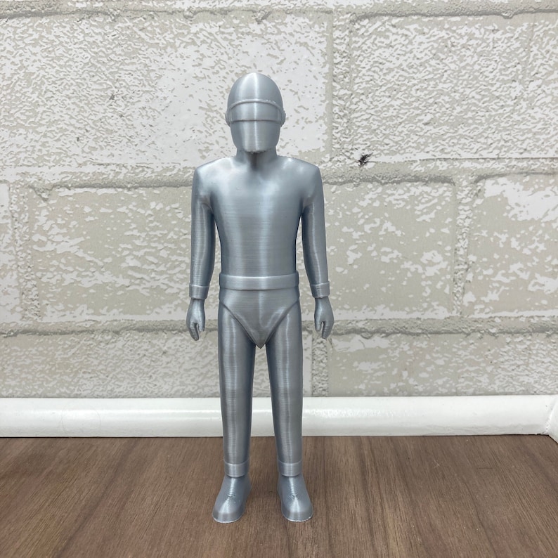May include: A silver-colored 3D printed figurine of a robot wearing a helmet. The robot has a human-like body with a smooth, metallic finish.