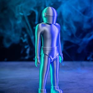 Op de afbeelding: Een zilveren 3D-geprint beeldje van een robot met een helm met vizier. De robot staat op een grijs oppervlak met een blauwe en groene rookachtergrond.