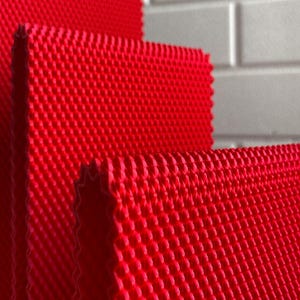 May include: Close-up of several layers of bright red, textured fabric. The fabric has a repeating pattern of small, raised squares, creating a grid-like appearance. The edges of the fabric are visible, showing a slightly jagged, cut edge.