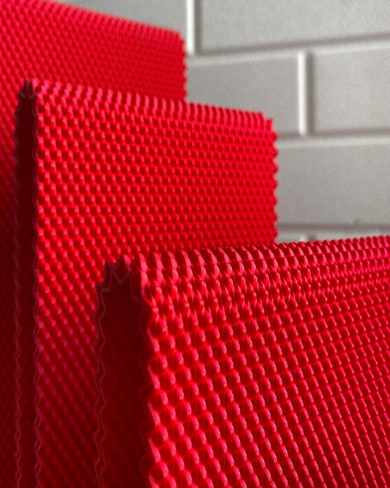 May include: Close-up of several layers of bright red, textured fabric. The fabric has a repeating pattern of small, raised squares, creating a grid-like appearance. The edges of the fabric are visible, showing a slightly jagged, cut edge.
