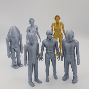 May include: A collection of six silver and gold robot figurines. The robots are standing on a white surface.