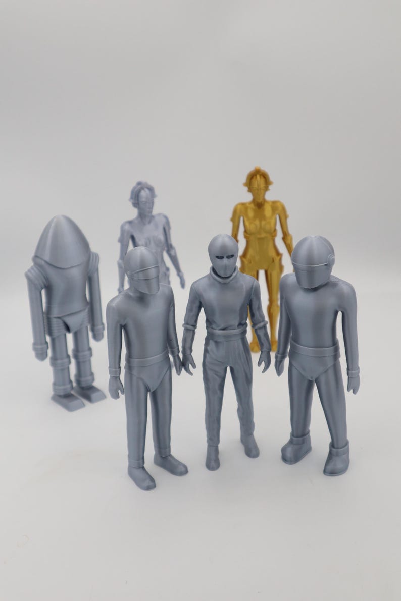 May include: A collection of six silver and gold robot figurines. The robots are standing on a white surface.