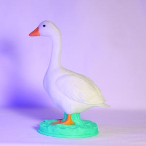 以下が含まれることがあります： オレンジ色のくちばしと足を持つ白いガチョウの置物。緑色の台座の上に立っています。ガチョウは横向きで、長い首をしています。これはおそらく装飾品です。
