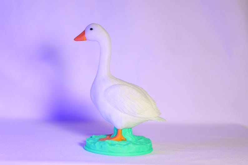 以下が含まれることがあります： オレンジ色のくちばしと足を持つ白いガチョウの置物。緑色の台座の上に立っています。ガチョウは横向きで、長い首をしています。これはおそらく装飾品です。
