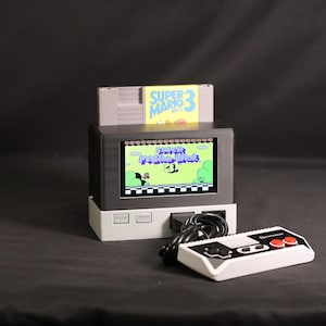 以下が含まれることがあります： 上部のスロットにスーパーマリオブラザーズ3のカートリッジが挿入された、白と黒のポータブルビデオゲーム機。画面にはスーパーマリオブラザーズのタイトル画面が表示されています。2つの赤いボタンが付いた長方形の白と黒のゲームコントローラーが接続されています。