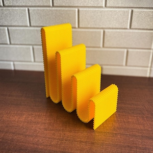 May include: Four bright yellow, textured, rectangular objects of varying sizes are arranged in a row. The objects have a folded, U-shaped design. The objects are on a dark brown surface, with a white brick wall in the background.