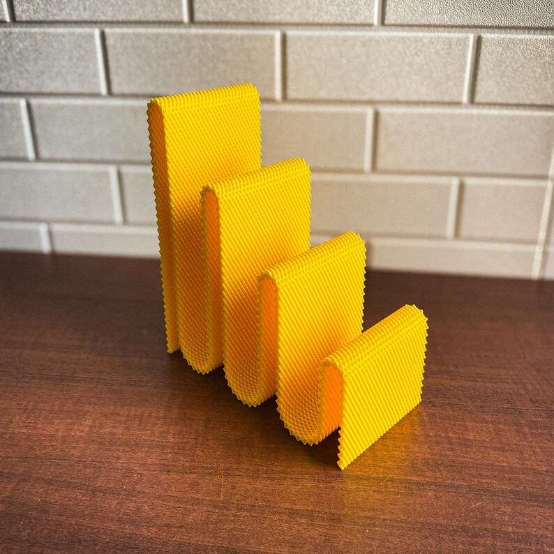 May include: Four bright yellow, textured, rectangular objects of varying sizes are arranged in a row. The objects have a folded, U-shaped design. The objects are on a dark brown surface, with a white brick wall in the background.