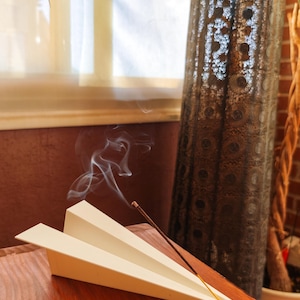 Op de afbeelding: Een witte, papieren vliegtuigvormige wierookhouder met een brandende wierookstok, die rook afgeeft. De houder staat op een houten oppervlak, met een decoratieve metalen lamp op de achtergrond. De scène straalt rust en ontspanning uit.