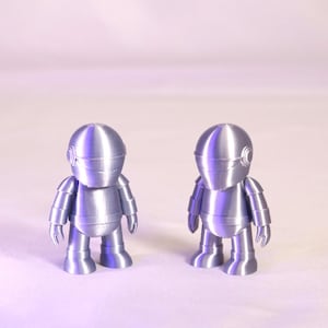 May include: Two silver-colored robot figurines. Each robot has a rounded helmet, segmented torso, and articulated limbs. The figures are standing upright, with a smooth, metallic finish. The background is a soft, gradient of light purple.