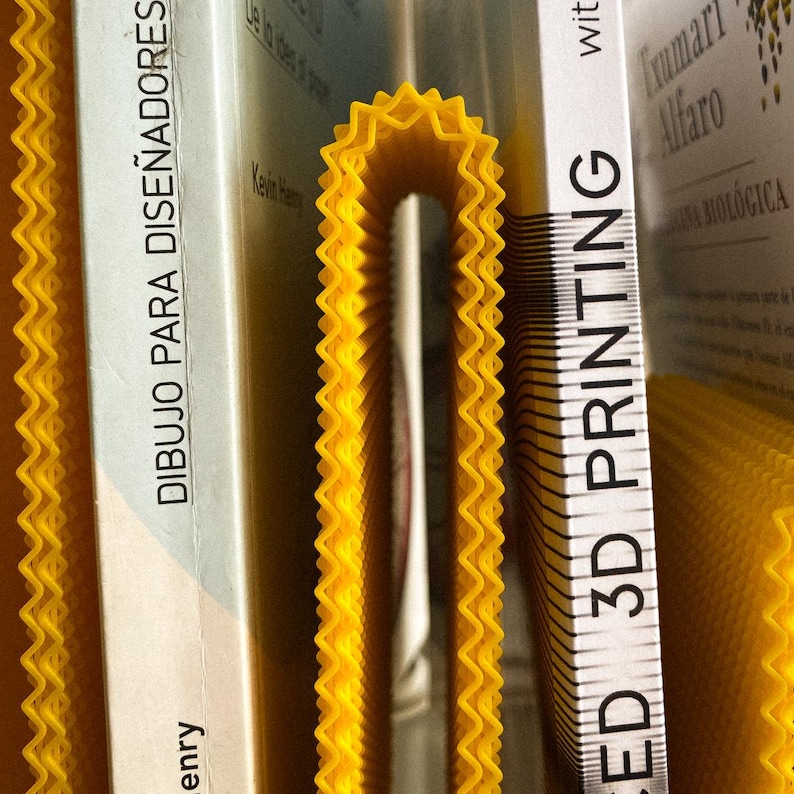 May include: A close-up of yellow, wavy, 3D-printed items, possibly for decoration or practical use. The items are placed near the spines of several books. One book spine reads "3D PRINTING". The items have a repeating, zig-zag pattern.
