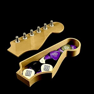 Puede incluir: Una caja de madera con forma de clavijero de guitarra llena de p&uacute;as. La caja est&aacute; abierta, revelando varias p&uacute;as de colores, incluyendo negras, blancas y moradas. Una forma separada de clavijero de madera con clavijas de afinaci&oacute;n se encuentra encima de la caja. El fondo es negro.