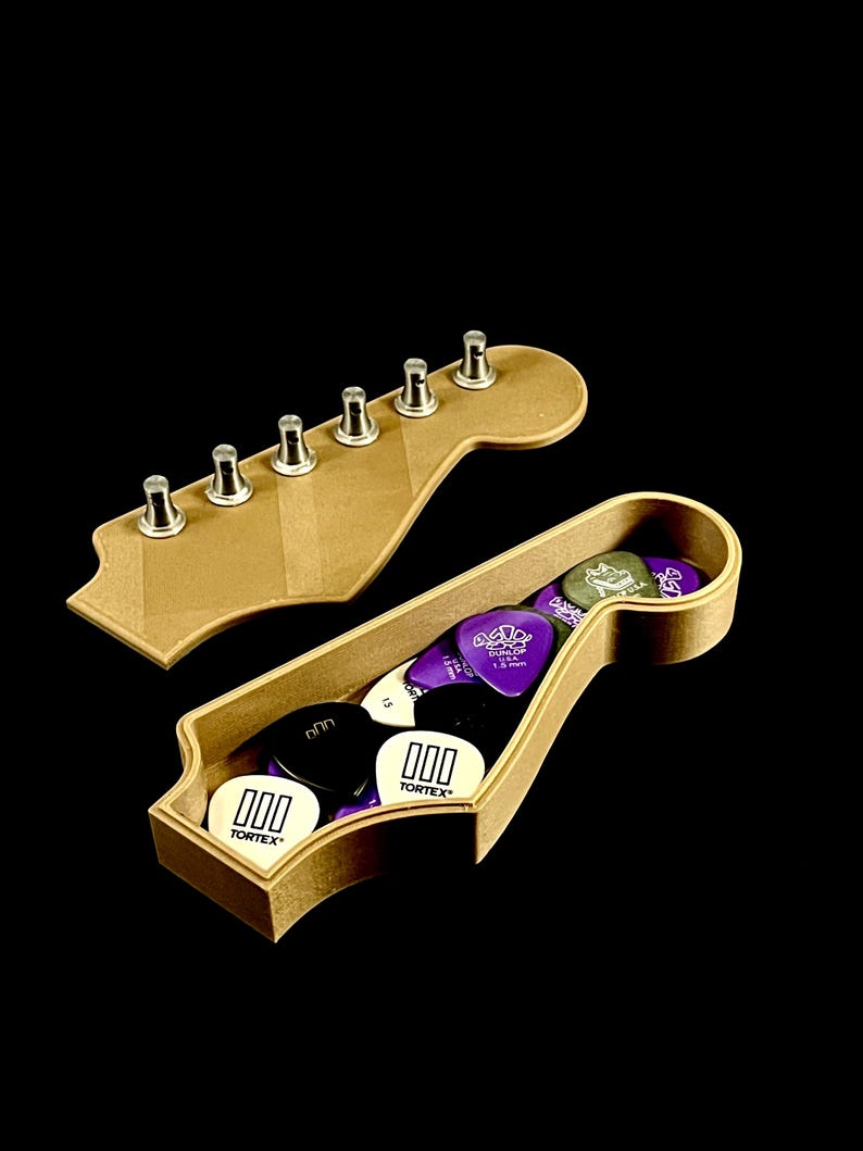 Puede incluir: Una caja de madera con forma de clavijero de guitarra llena de p&uacute;as. La caja est&aacute; abierta, revelando varias p&uacute;as de colores, incluyendo negras, blancas y moradas. Una forma separada de clavijero de madera con clavijas de afinaci&oacute;n se encuentra encima de la caja. El fondo es negro.