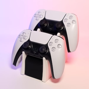 May include: Two white wireless game controllers with black buttons and a black charging stand. The controllers are on a pink and purple gradient background.