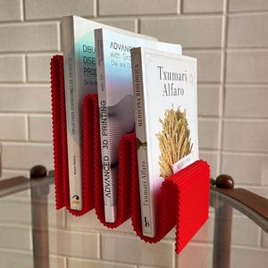 May include: A red, concertina-style book holder with three books. The books have white covers and titles including "DIBUJO PARA DISE&Ntilde;ADORES DE PRODUCTO", "ADVANCED 3D PRINTING", and "MEDICINA BIOL&Oacute;GICA". The holder is mounted on a wall.