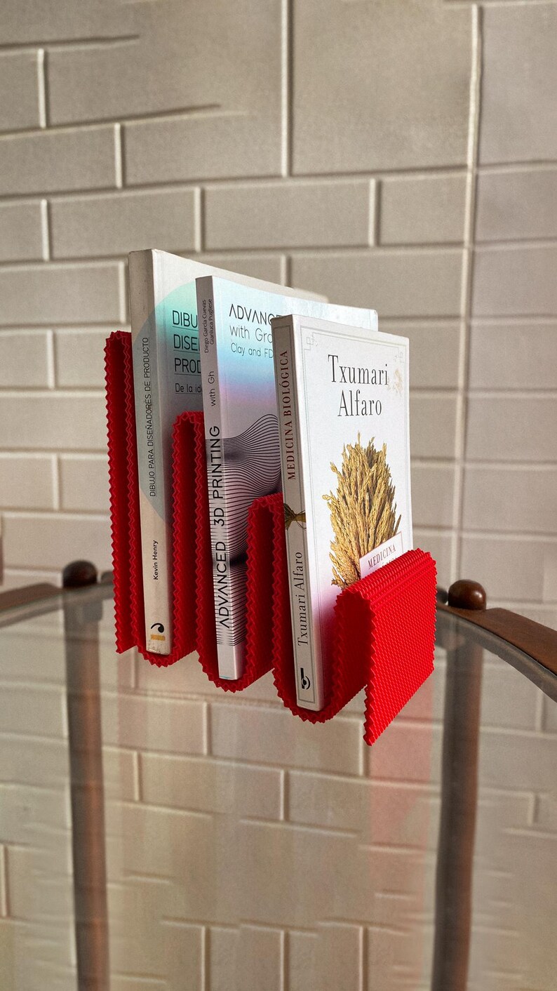 May include: A red, concertina-style book holder with three books. The books have white covers and titles including "DIBUJO PARA DISE&Ntilde;ADORES DE PRODUCTO", "ADVANCED 3D PRINTING", and "MEDICINA BIOL&Oacute;GICA". The holder is mounted on a wall.
