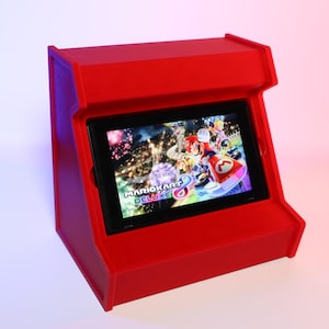 Pode incluir: Um gabinete de arcade vermelho impresso em 3D para um console Nintendo Switch. O gabinete tem uma ranhura para o console e uma tela. A tela mostra o jogo Mario Kart 8 Deluxe.