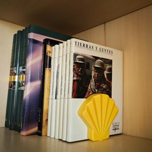 May include: A collection of books on a wooden shelf, including titles like "TIERRAS Y GENTES" and "EGIPTO." A bright yellow seashell-shaped bookend is placed next to the books. The books have various colors and sizes.