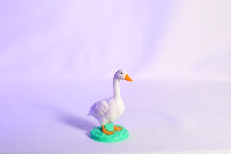 May include: A white goose figurine with an orange beak and feet, standing on a green base. The figurine is set against a soft purple background, creating a whimsical and charming aesthetic.