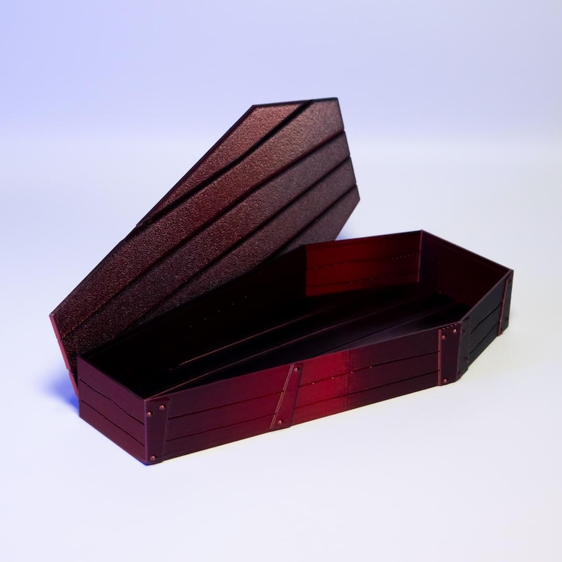 Puede incluir: Un ata&uacute;d rojo oscuro abierto, con tapa a juego. El ata&uacute;d est&aacute; hecho de tablones de madera y tiene detalles met&aacute;licos. La tapa est&aacute; ligeramente entreabierta, revelando el interior. Un art&iacute;culo decorativo &uacute;nico.