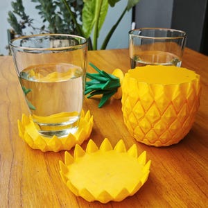 Könnte beinhalten: Vier gelbe Untersetzer in Ananasform, auf einem steht ein Glas Wasser. Die Untersetzer sind aus einem strukturierten Material gefertigt und haben ein realistisches Ananas-Design.