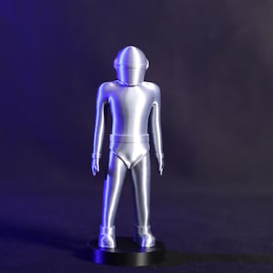 May include: A silver-coloured robot figurine with a helmet and a black base. The robot is standing with its arms at its sides.