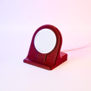 Könnte beinhalten: Ein roter kabelloser Ladeständer für eine Smartwatch. Der Ständer hat einen kreisförmigen Ladebereich und eine strukturierte Oberfläche.