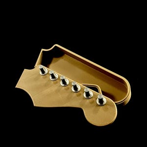 Puede incluir: Una caja en forma de clavijero de guitarra marr&oacute;n claro con seis clavijas plateadas. La caja est&aacute; abierta, revelando un interior a juego. El fondo es negro s&oacute;lido.