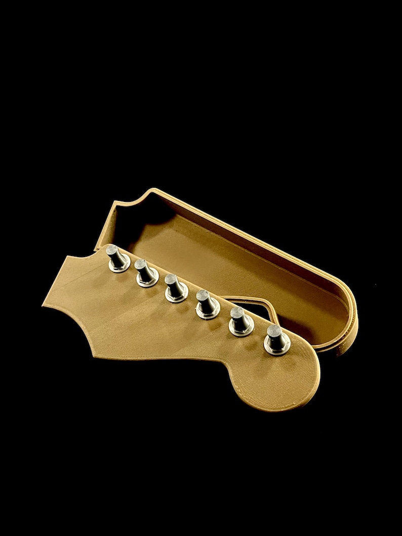 Puede incluir: Una caja en forma de clavijero de guitarra marr&oacute;n claro con seis clavijas plateadas. La caja est&aacute; abierta, revelando un interior a juego. El fondo es negro s&oacute;lido.
