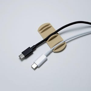May include: A beige cable organizer holding a black and a white USB-C cable. The organizer has three curved slots to hold the cables. The cables have black and white connectors.