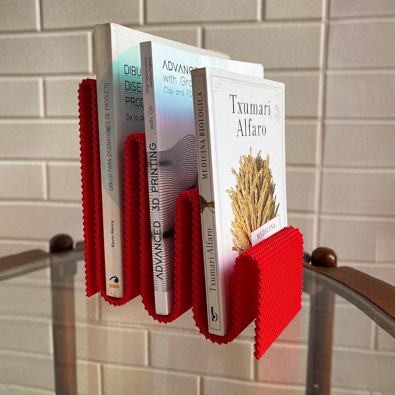 May include: A red, concertina-style book holder with three books. The books have white covers and titles such as "Advanced 3D Printing" and "Medicina Biologica." The holder is made of a textured material and is wall-mounted.
