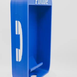 May include: A blue rectangular wall-mounted phone holder with rounded corners. The top features the white text "Phone" and the side displays a white telephone handset icon. The interior includes a small horizontal ledge and a circular hole at the base.