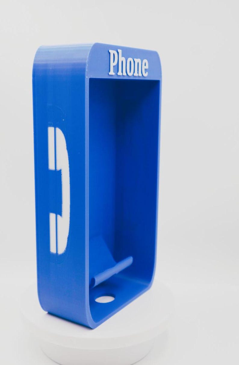 May include: A blue rectangular wall-mounted phone holder with rounded corners. The top features the white text "Phone" and the side displays a white telephone handset icon. The interior includes a small horizontal ledge and a circular hole at the base.