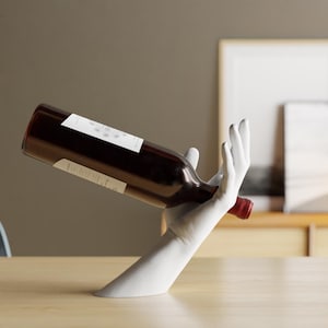 Könnte beinhalten: Weiße Keramik-Handskulptur, die eine Flasche Rotwein hält. Die Hand ist so positioniert, als würde sie den Wein einschenken. Die Flasche ruht auf der Handfläche.