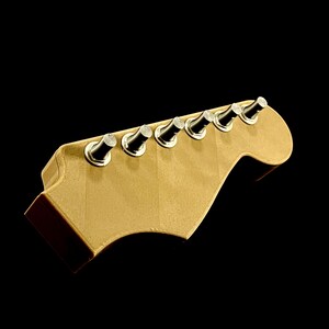 Puede incluir: Un clavijero de guitarra con seis clavijas. El clavijero es de color beige claro, con un borde marr&oacute;n oscuro. Las clavijas son plateadas y negras. El fondo es negro.