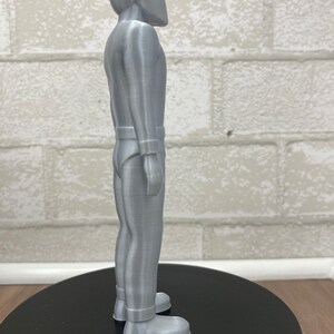 May include: A silver-colored 3D printed figurine of a person wearing a helmet and a jumpsuit. The figurine is standing on a black circular base.