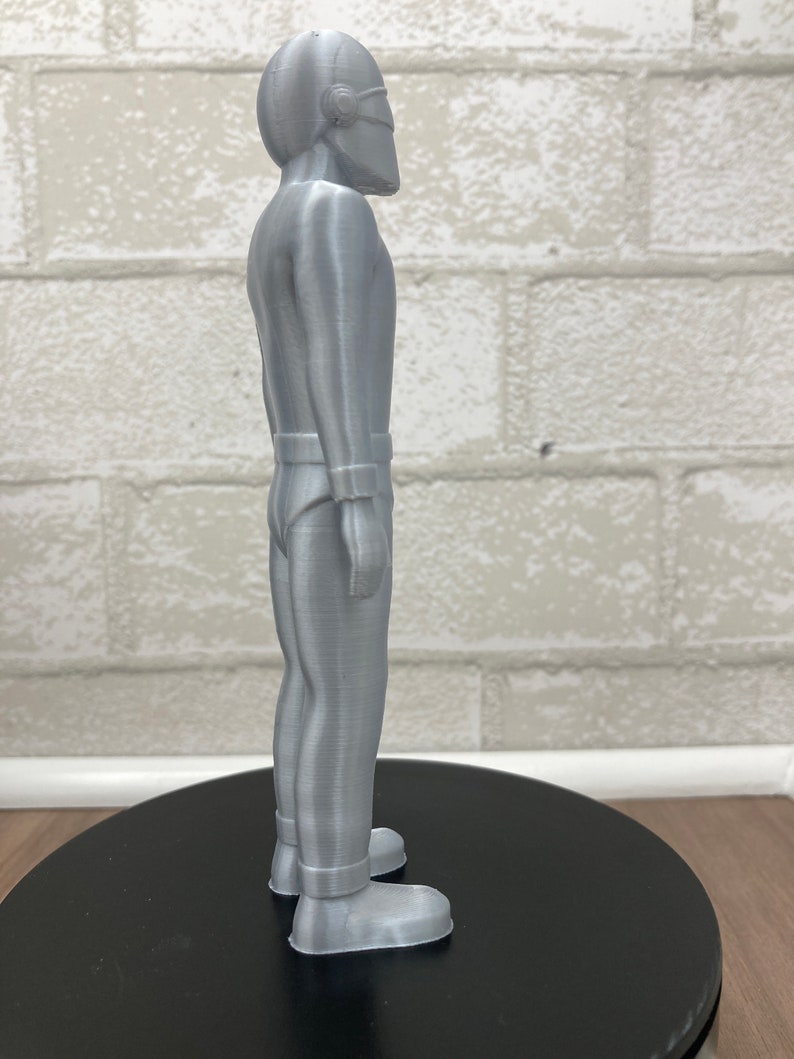 May include: A silver-colored 3D printed figurine of a person wearing a helmet and a jumpsuit. The figurine is standing on a black circular base.