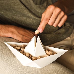 Peut inclure: Un bateau en papier blanc rempli de pistaches. Une main tend la main vers le bateau, tenant une seule pistache. Le bateau est posé sur une surface de couleur claire, avec une personne portant une chemise verte en arrière-plan.