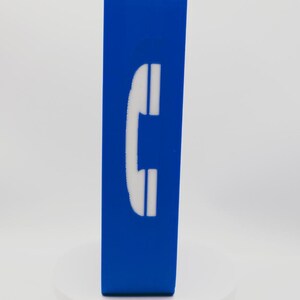 May include: A rectangular blue block featuring a white telephone handset icon centered on the front face. The object stands upright on a circular white base, displaying a smooth, solid-colored surface with a clear, high-contrast graphic symbol.
